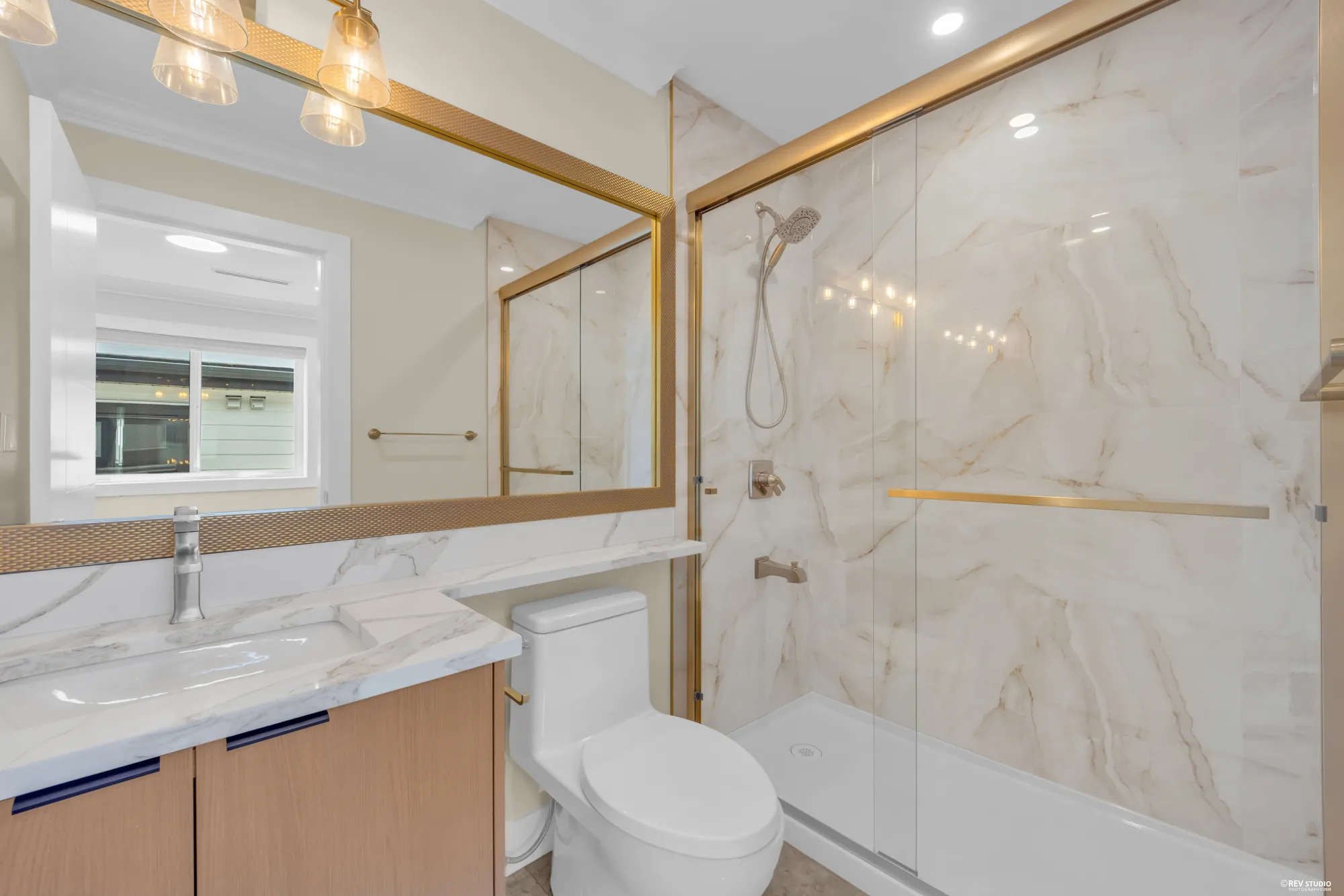 A bathroom with a tub and shower in marbled tile behind glass, brass fittings