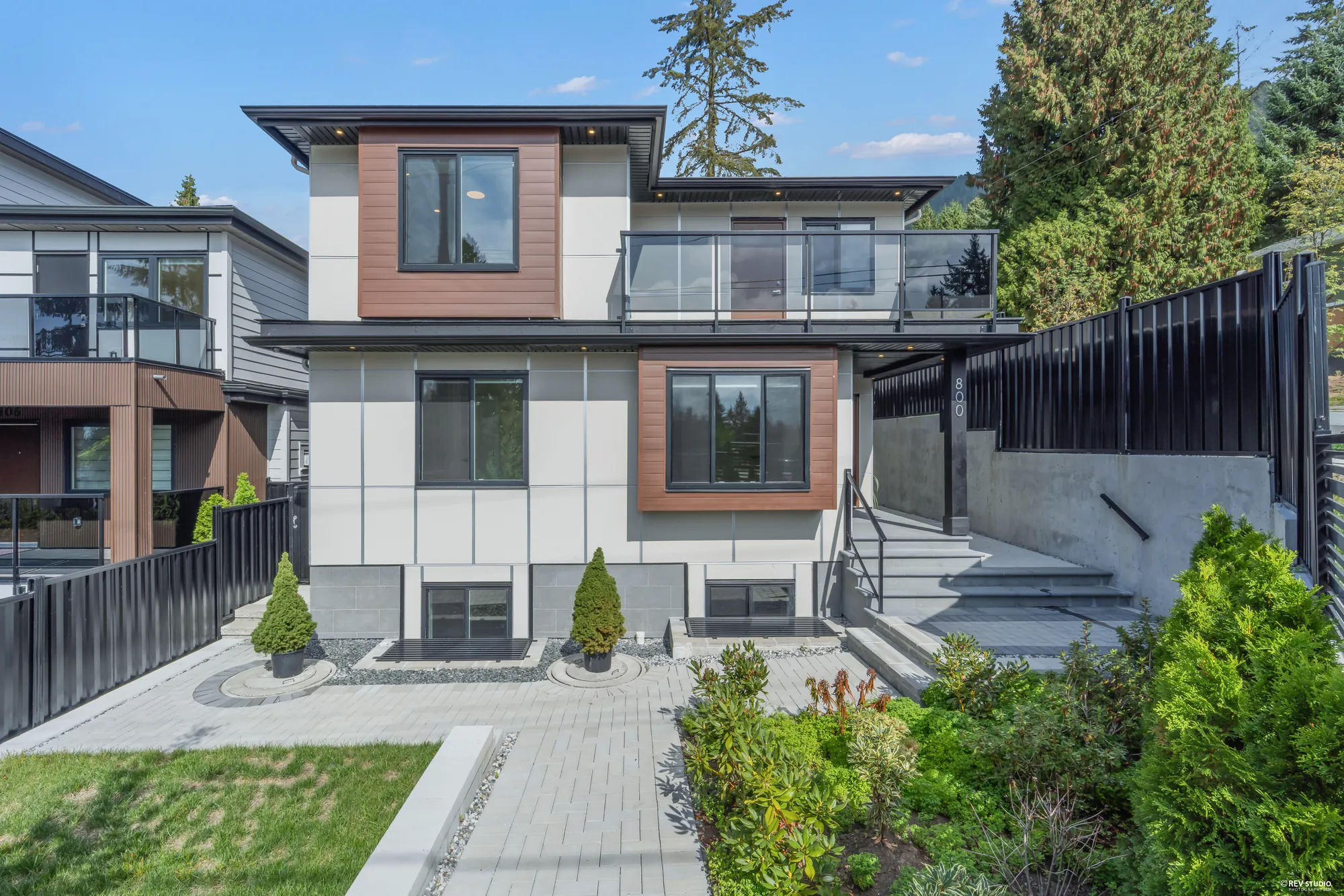 The front of the house in daylight: three levels of white and grey panels with wood-look accents, the balcony over the entry, planters and turf inside a black fence