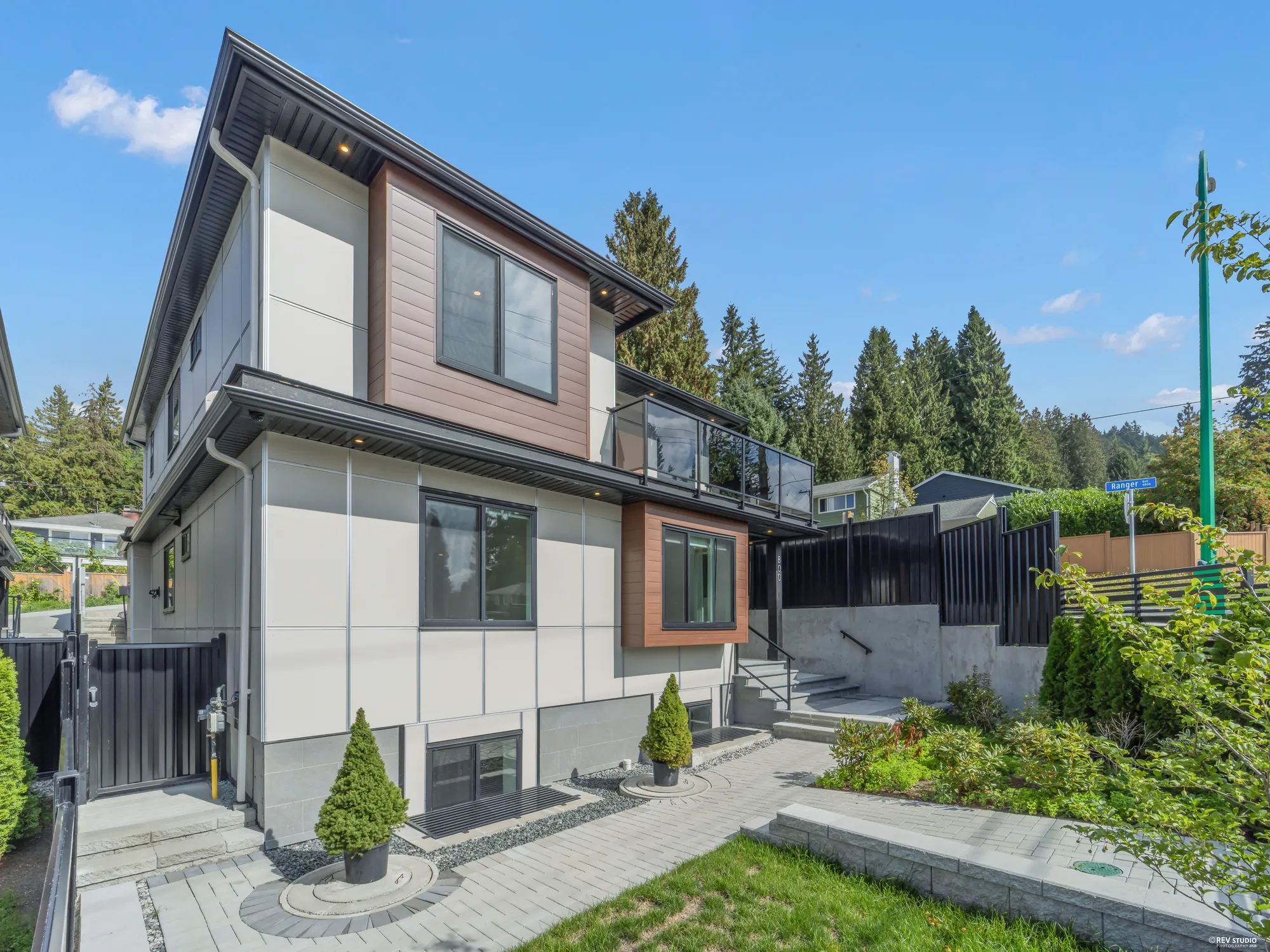 The house from the side lawn: the wood-panelled bays, the glass balcony above, small conifers in planters by the black fence