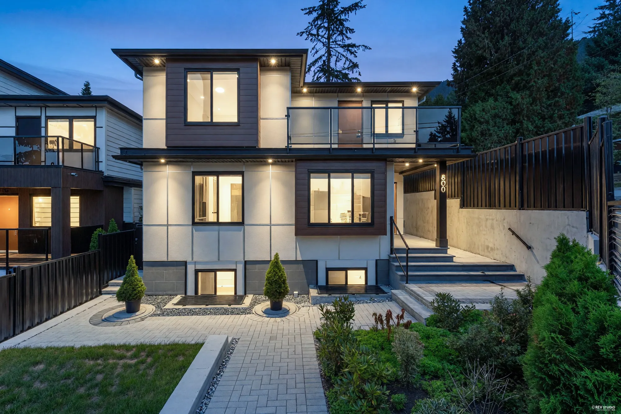 800 Montroyal Boulevard from the street at dusk: three levels of white and grey panels with wood-look accents, every window lit, the black fence and planters in front