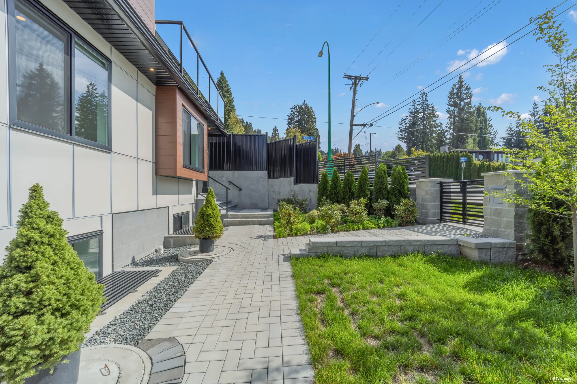 The paved path and turf in front of the house, conifers in planters, the stair up beside the house, the black fence and the street beyond