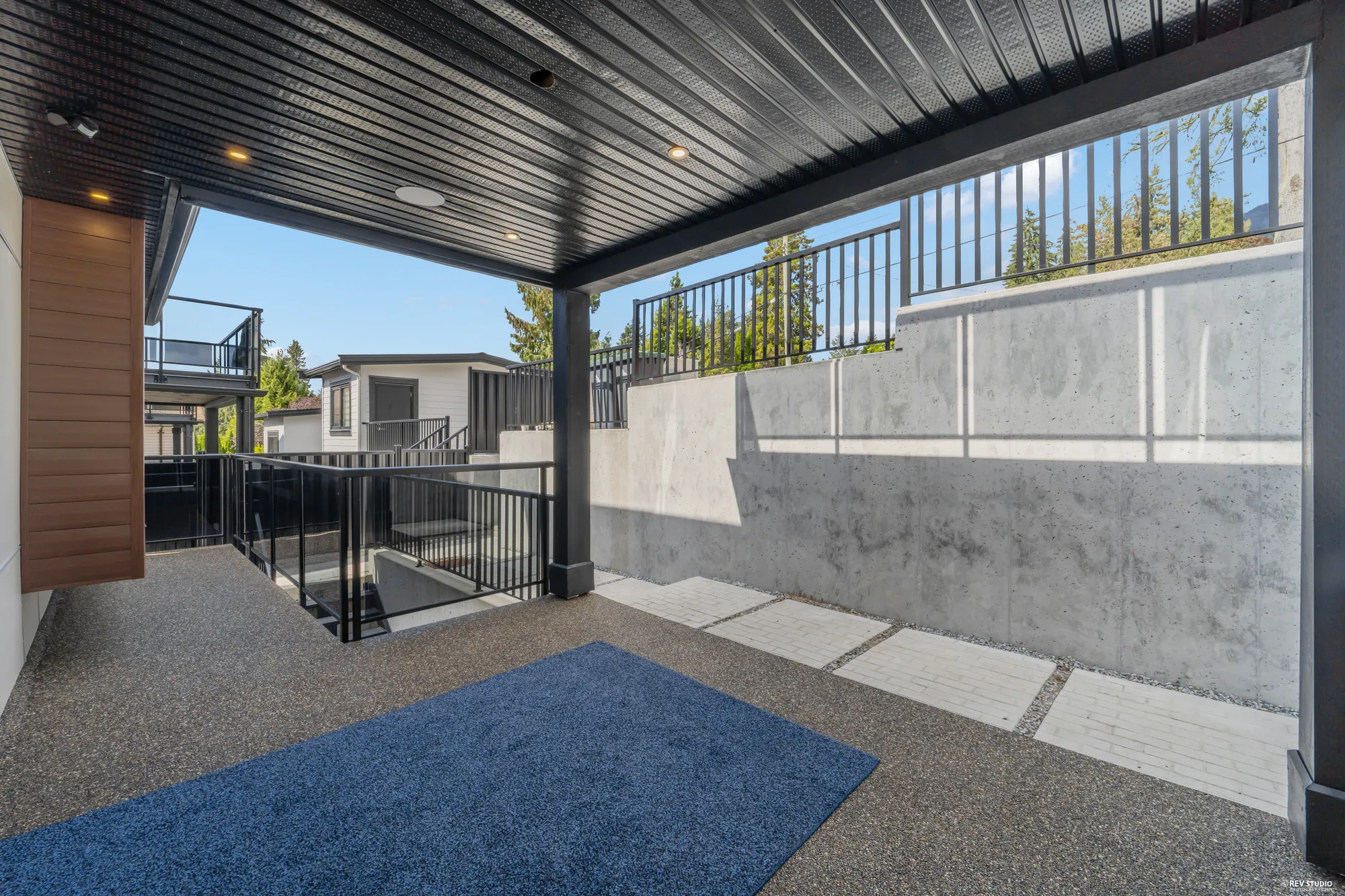 The covered patio under the deck: a black soffit, concrete walls, a blue mat, a picket rail to the stair