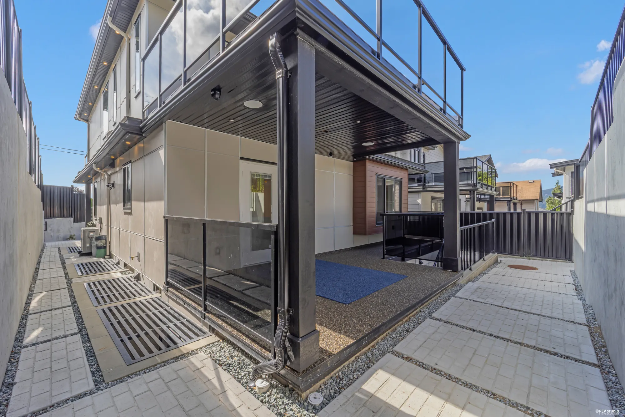 The side passage in pavers and gravel, the covered patio under the deck with its black soffit and a blue mat